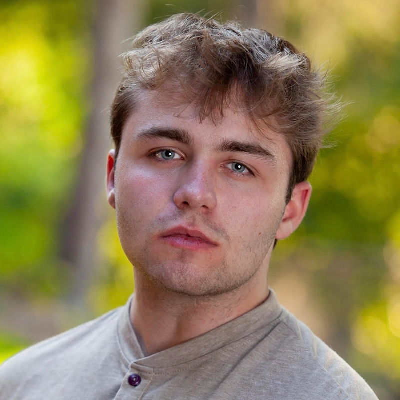 A young man, Timothy Thomas, with tousled light brown hair and blue eyes stares at the camera with a neutral expression. He wears a beige buttoned shirt. The background is softly blurred with green and yellow tones, suggesting an outdoor setting.