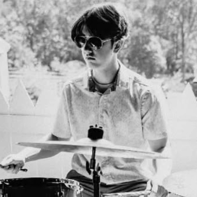 A young person wearing sunglasses and a short-sleeved shirt plays a drum set outdoors. Sunlight filters through trees behind William Nicoll, with a white picket fence in view. The image is in black and white.