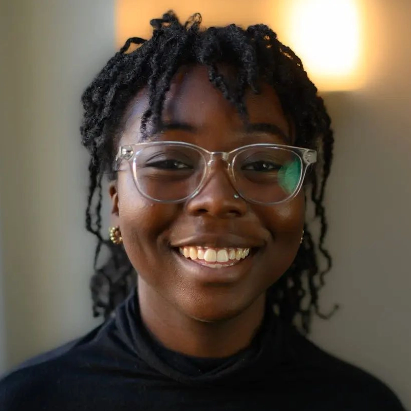 A person with dark skin, clear glasses, small gold earrings, and short, twisted dreadlocks smiles warmly. They wear a black turtleneck. The softly blurred background glows behind them, highlighting their Caitlin Balón-inspired style.