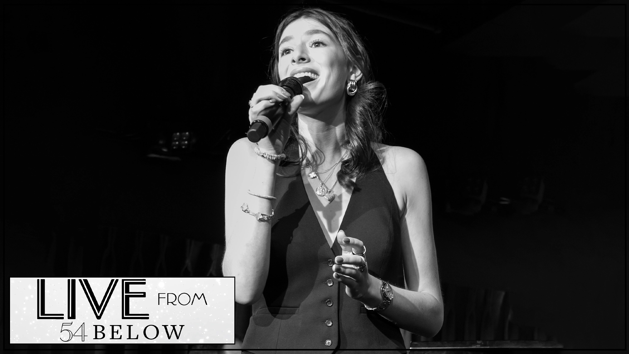 A young woman sings passionately on stage with a microphone in hand, dressed in a sleeveless top and jewelry. The scene is in black and white. In the corner, a sign reads “LIVE FROM 54 BELOW.” The background is dimly lit, focusing attention on the performer.