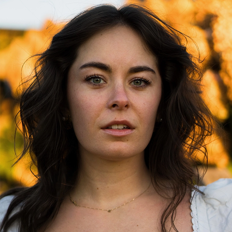 Claire Leyden, a woman with wavy brown hair and green eyes, looks directly at the camera. She wears a white top and a delicate gold necklace. The softly blurred background glows with warm, golden light and hints of foliage. Her expression is calm and neutral.