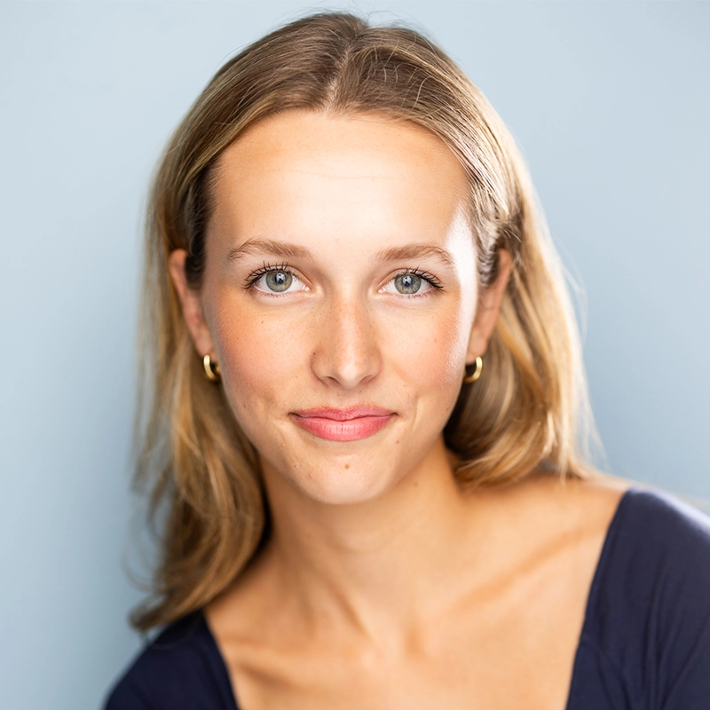 A young woman, Addison Wivagg, with long blonde hair, blue eyes, and light skin smiles gently at the camera. She wears small gold hoop earrings and a navy blue top, set against a soft light blue background with bright, even lighting.