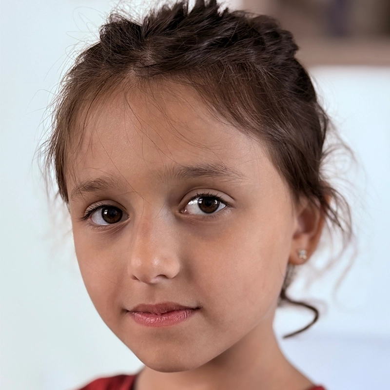 A young girl, Olympia Eyal, with brown eyes and long brown hair tied back looks directly at the camera. She has light skin, small stud earrings, and wears a red top. The background is softly blurred, emphasizing her gentle expression.