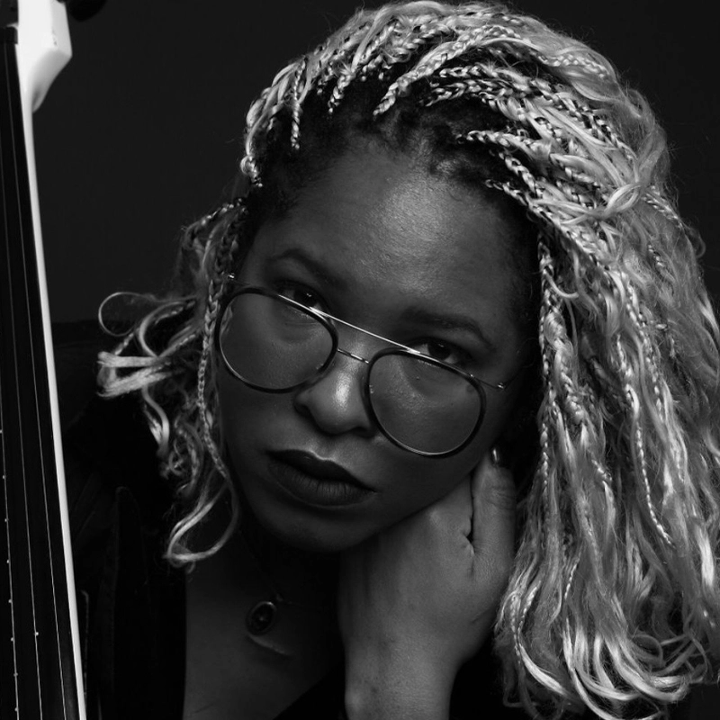Black and white close-up of Sarah Overton with long, light-colored braided hair, wearing large round glasses. She has a serious expression, resting her chin on her hand. Part of a stringed instrument appears on the left against a dark background.