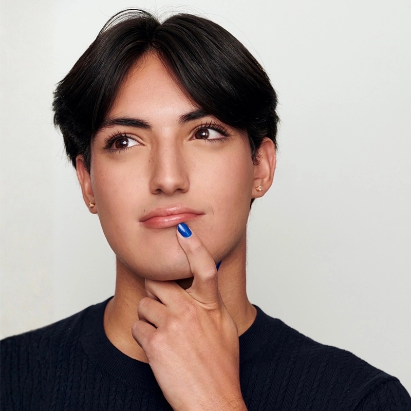 Sophia Leverett, with short dark hair and light skin, looks thoughtful, touching her chin with a finger painted blue. She wears a dark sweater and small earrings, posing against a plain light background, her gaze directed up and to the side.