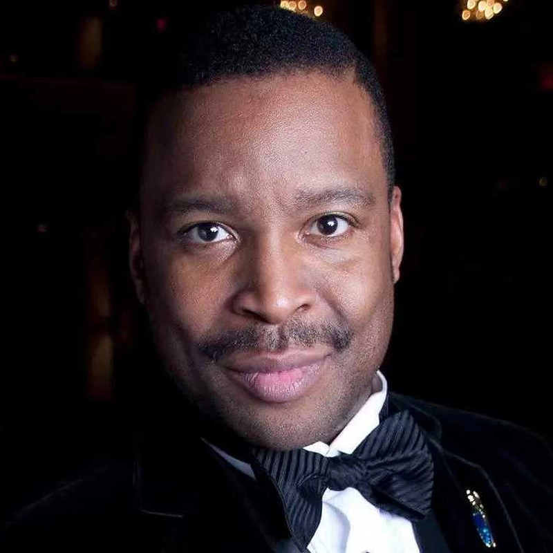 Chauncey Packer, in formal attire including a black tuxedo, bow tie, and white shirt, is smiling slightly and looking directly at the camera. The dark background with blurred yellow lights highlights his face.