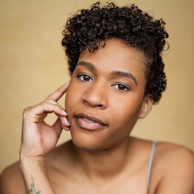 Alice K. Tinari, with short, curly dark hair and warm brown skin, looks calmly at the camera. Wearing a thin strap top, she gently touches her face, revealing a small green vine tattoo on her wrist against a soft beige background.