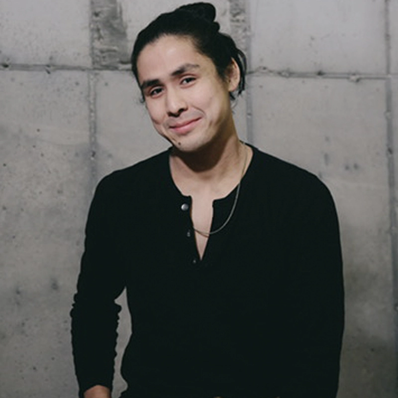 Chris Peters, with long dark hair in a bun, wearing a black henley shirt and a thin chain necklace, stands against a gray concrete wall, looking at the camera with a gentle smile and a relaxed posture.