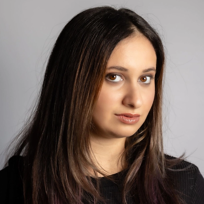 A young woman with long, straight brown hair and brown eyes looks at the camera. She is wearing a black top and has a neutral expression. The background is plain light gray, giving the portrait of Phebe Barnett a calm and understated tone.