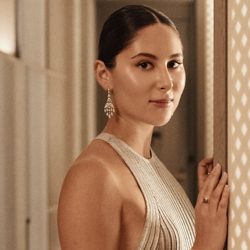 A woman with dark hair pulled back, resembling Eden Plepler, wears a sleeveless silver pleated dress and dangling earrings. She stands indoors, facing the camera with a soft smile, one hand resting on a patterned wall as warm lighting highlights her elegance.