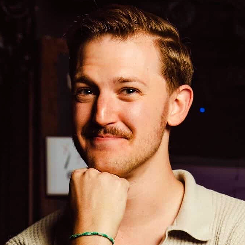 A smiling man, resembling Edward Precht, with light brown hair and a mustache rests his chin on his fist. He wears a beige collared shirt and a green bracelet. The dim background draws attention to his friendly expression.