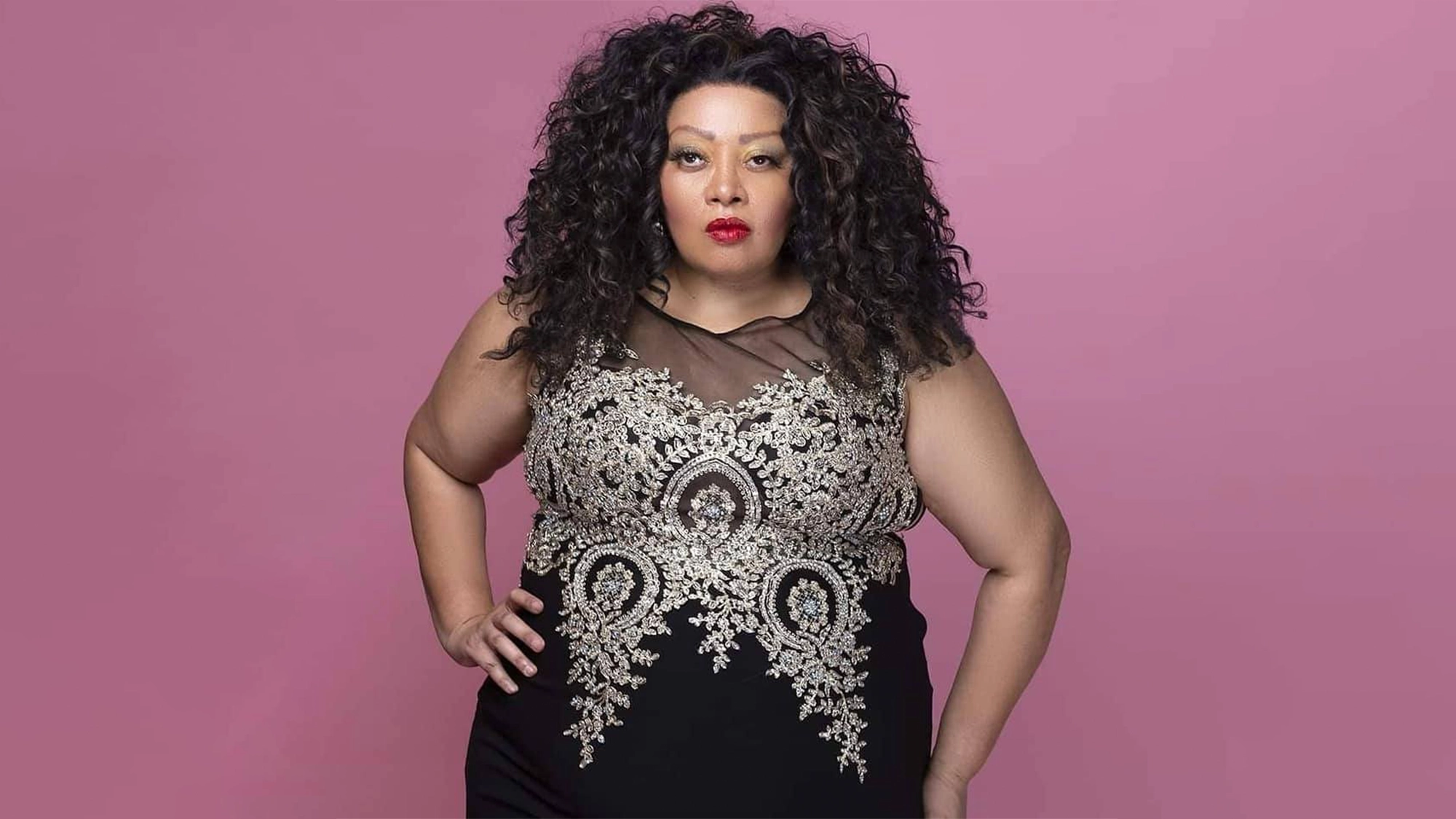 Queen Diva site image 26 A woman with long, curly dark hair stands confidently against a pink background, embodying the strength of a woman. In a sleeveless black dress with gold embroidery and bold red lipstick, she channels a true Queen Diva—an elegant Phyllis Hyman tribute.