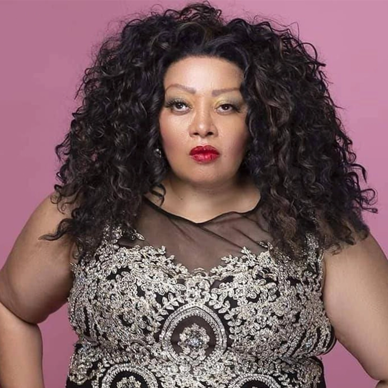 A confident woman with long, voluminous curly black hair poses against a pink background. She wears a sleeveless black dress with intricate silver embroidery and sheer mesh at the top, bold red lipstick, and subtle eye makeup.