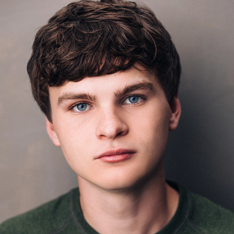 A young person, Braxton Quinney, with short, wavy brown hair and striking blue eyes looks seriously at the camera. They have fair skin and wear a dark green top, with a neutral, softly blurred background that keeps the focus on their face.
