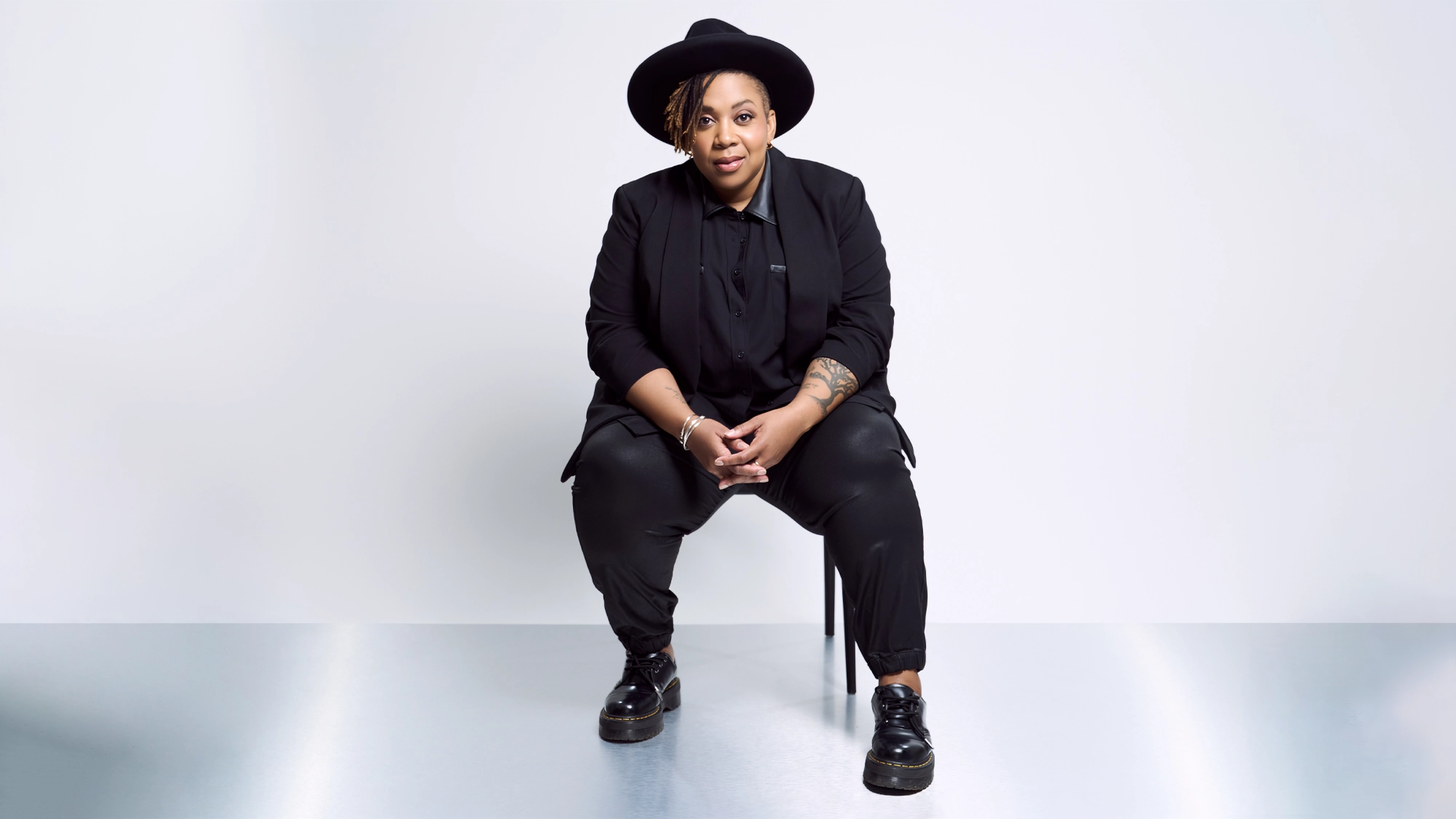 A person with short, curly hair sits on a stool against a plain white background, dressed in REAL Black style—black wide-brim hat, suit, shirt, and shoes. Crystal Monee Hall's Juneteenth Celebration inspires their confident pose and tattooed forearm.