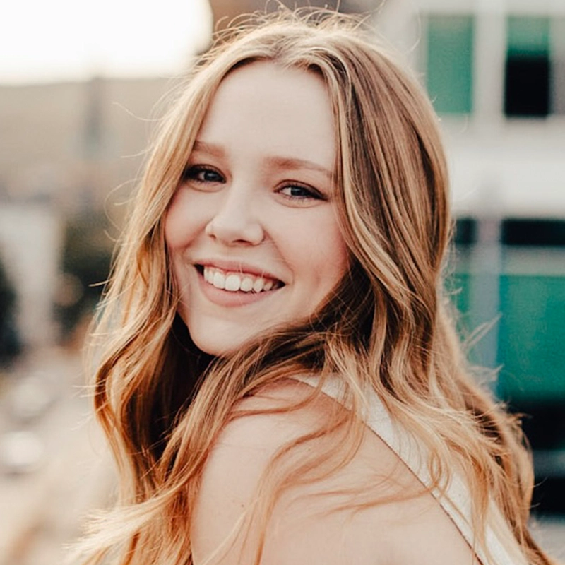 A young woman with long, wavy blonde hair smiles brightly at the camera. She is outdoors, with blurred buildings and a soft, warm light in the background. She wears a sleeveless light top and appears happy and relaxed.