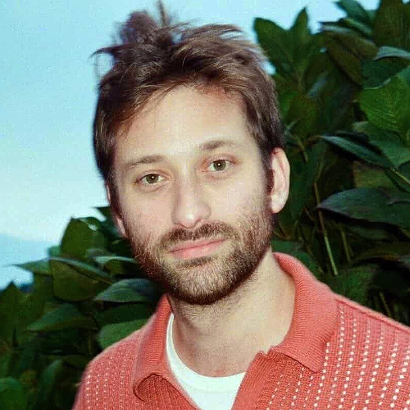 A man with light skin, short tousled brown hair, and a beard wears a coral polo shirt with a white undershirt. He stands outdoors in front of large green leaves, with a soft blue sky in the background, looking at the camera.