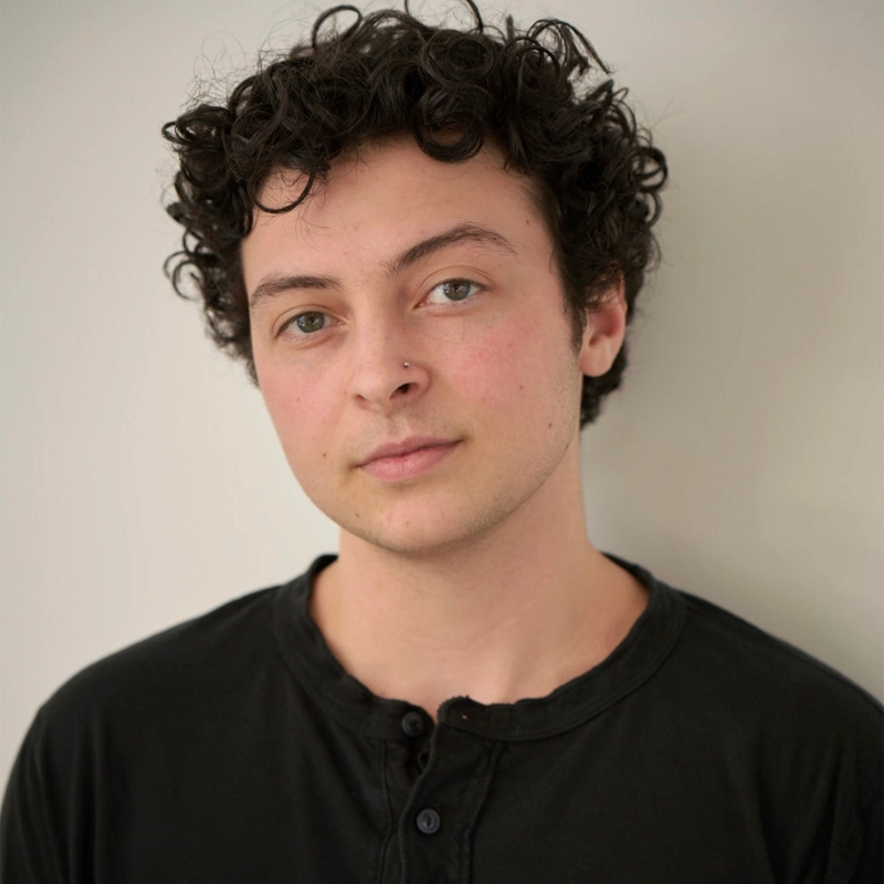 A young person with short, curly dark hair and light skin stands against a plain off-white background. They have a slight smile, green eyes, and a nose piercing. They wear a black, buttoned shirt and look directly at the camera with a relaxed expression.