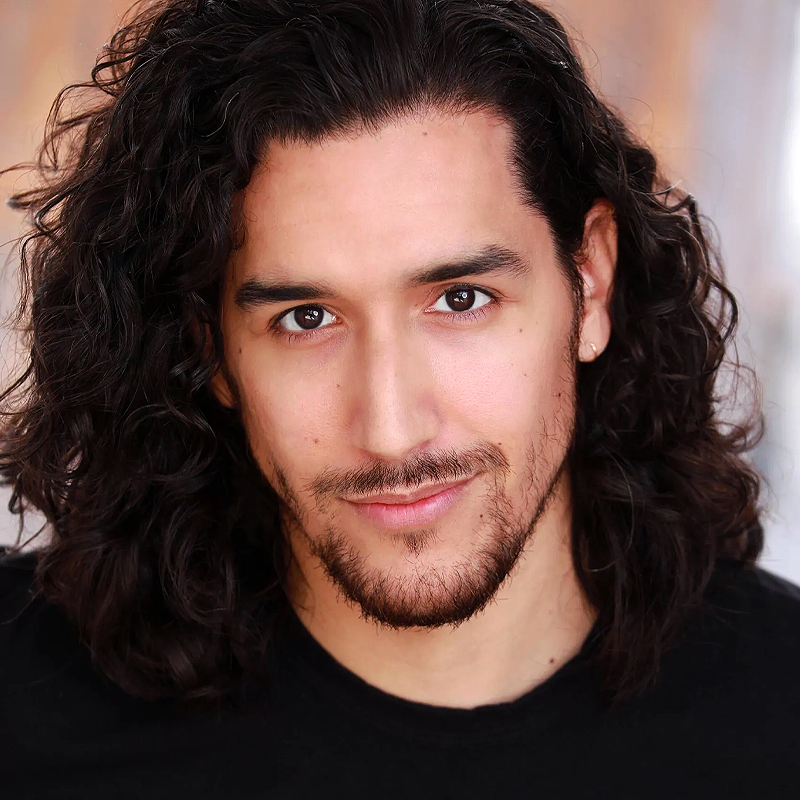 Christopher M. Ramirez, a man with long, curly dark hair and a trimmed beard, looks directly at the camera, smiling slightly. He has light skin and brown eyes, wears a black shirt, and is set against a softly blurred warm brown and gray background.