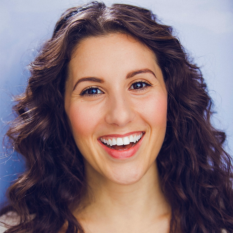 A young woman with long, wavy brown hair smiles brightly at the camera. She has clear skin, rosy cheeks, and is wearing natural makeup. The background is a soft blue, and the lighting highlights her cheerful expression.