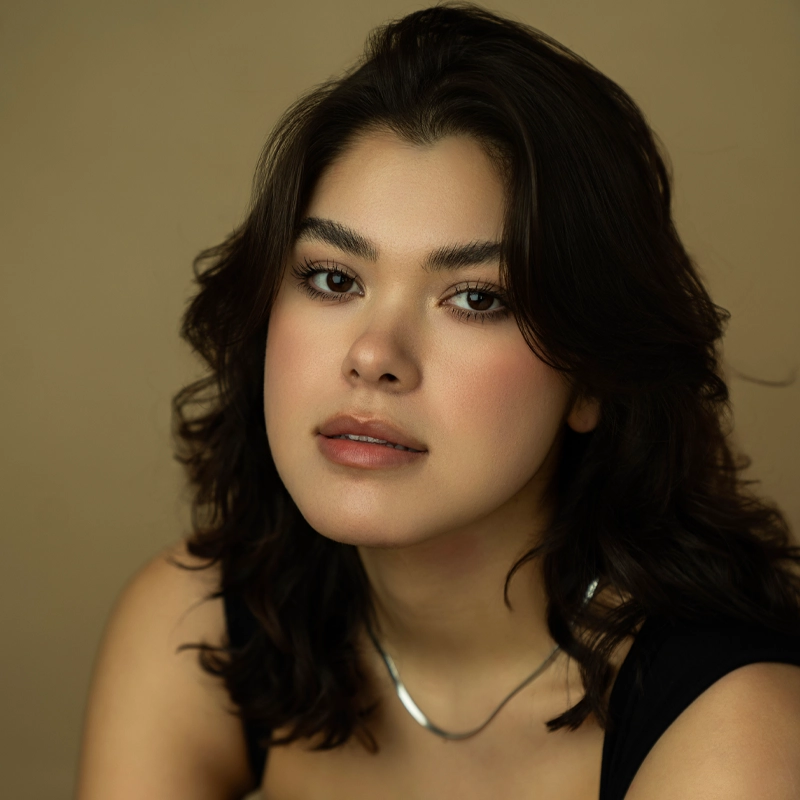 A young woman, Emma Kayden Reed, with wavy dark hair and natural makeup looks at the camera with a calm expression. She wears a black top and a silver necklace. The plain beige background creates a soft and warm atmosphere.