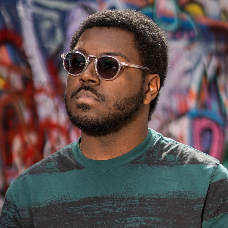 A man with short curly hair, a beard, and mustache wears round sunglasses and a green patterned shirt. Koleby Boyston stands outdoors in front of a colorful graffiti wall, looking slightly upward with a neutral expression.
