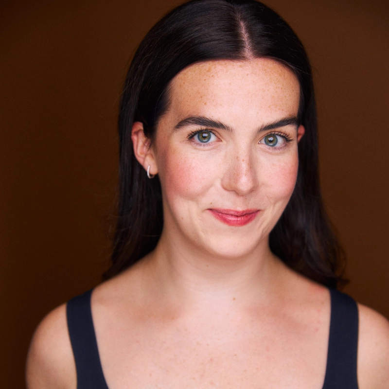 Meghan Rooney, a woman with long dark hair and fair skin, smiles softly at the camera. She has blue-green eyes, freckles, and wears a sleeveless dark top with small silver hoop earrings. The background is a solid warm brown.