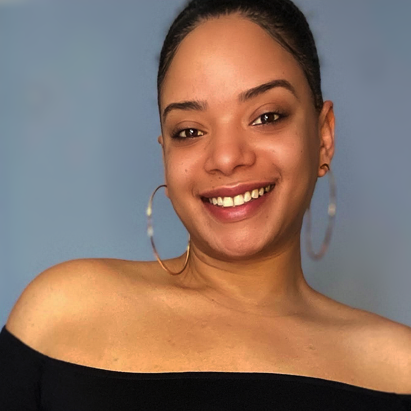 Lindsay Rose, with medium skin tone, smiles at the camera. She has dark hair pulled back, wears large hoop earrings, and an off-the-shoulder black top against a plain, light blue wall.