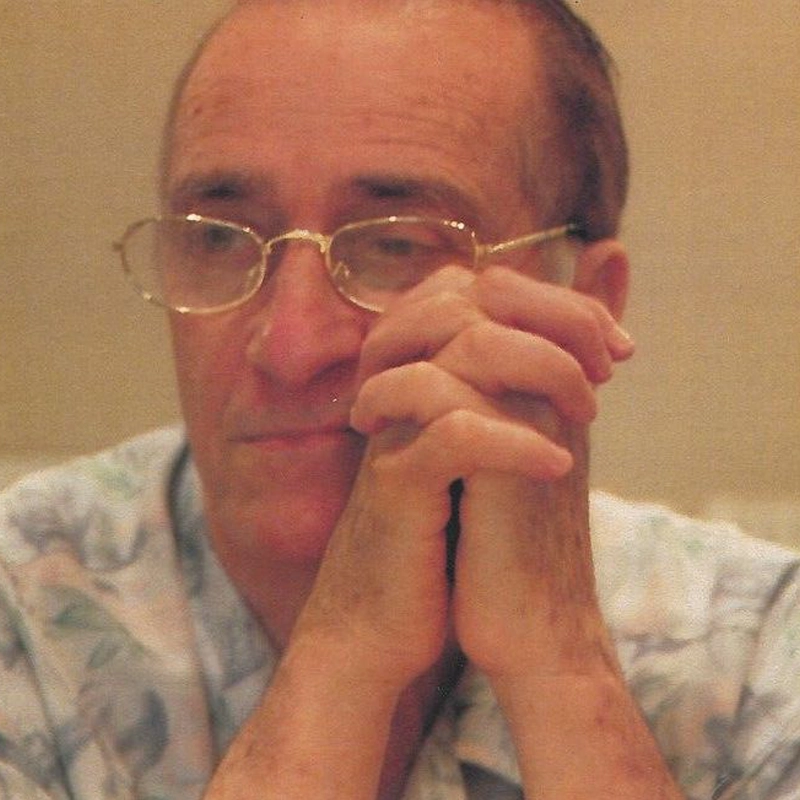 David Rothenberg, an older man with thinning hair and glasses, clasps his hands and rests his chin on them. He wears a light patterned shirt and looks thoughtful, gazing slightly downward against a warmly blurred background.