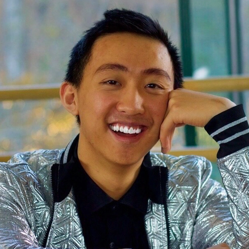 A smiling young man with short black hair sits indoors, resting his head on his hand. He wears a shiny silver geometric-patterned jacket over a black shirt. Soft natural light and blurred outdoor scenery are visible in the background.