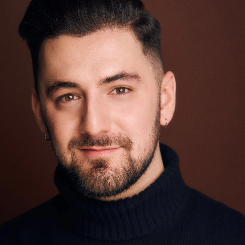 Alec Michael Ryan, a man with short dark hair and a trimmed beard, smiles softly at the camera. He wears a black turtleneck sweater and small hoop earrings. The solid brown background creates a warm, neutral setting for the portrait.