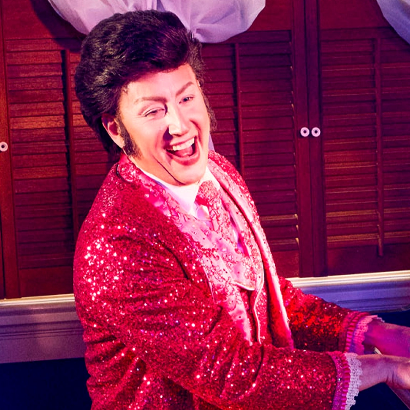 David Saffert, in a sparkling bright pink sequin jacket and ruffled shirt, smiles widely while playing the piano. The lighting is pinkish-purple, wood shutter doors are in the background, and his styled dark hair adds to his expressive, joyful vibe.