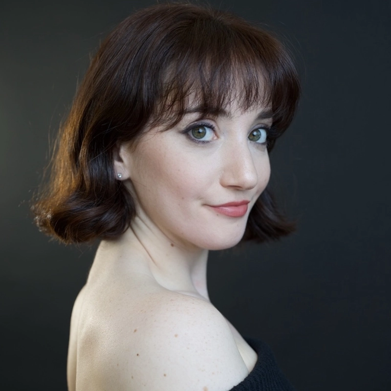A woman with fair skin and short, wavy brown hair looks over her shoulder at the camera. She has green eyes, subtle makeup, and wears a black off-the-shoulder top. The background is dark and plain, highlighting her face and shoulders.