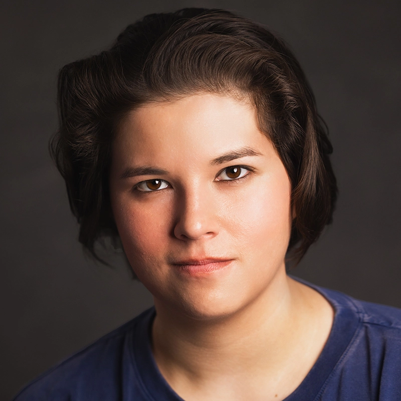 A person with short, dark brown hair and fair skin looks directly at the camera with a neutral expression. Maggie Weller is wearing a blue shirt, and the background is a plain, dark gray. Soft lighting highlights her face.
