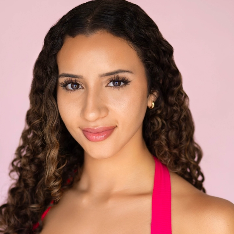 Shayla Lopez, a young woman with long, curly brown hair and glowing skin, smiles softly. She wears hoop earrings and a bright pink halter top against a smooth, light pink background, creating a warm and inviting atmosphere.