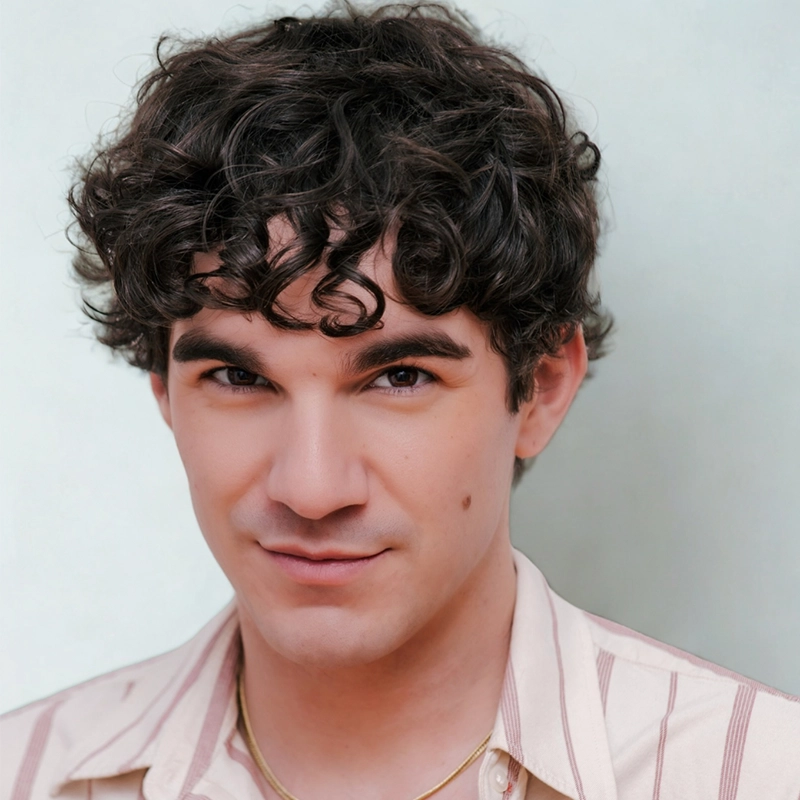 A young man with short, curly brown hair and fair skin looks at the camera with a slight smile. He has thick eyebrows, brown eyes, and a small mole on his cheek, wearing a light pink striped shirt and thin gold necklace—Maggie Weller style.
