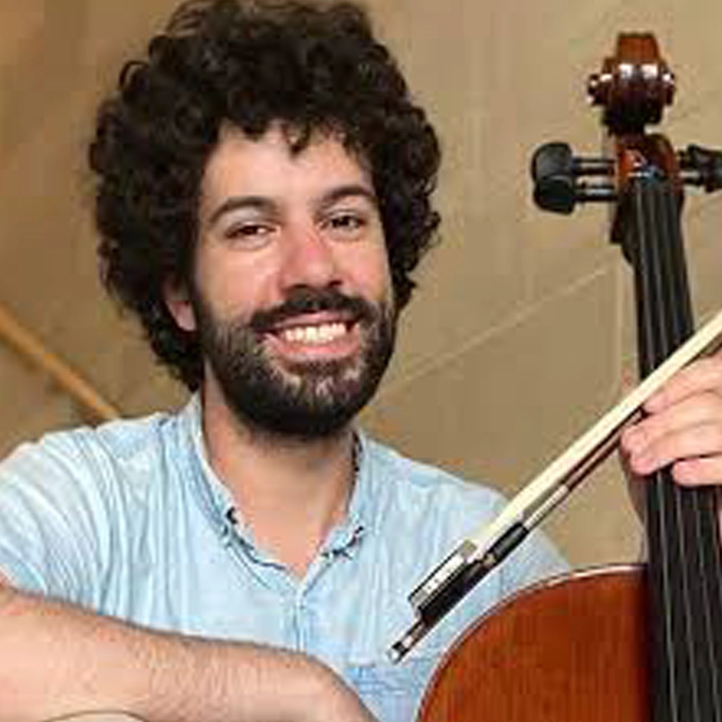 A smiling man with curly dark hair and a beard, identified as Chris Santos, wears a light blue shirt and holds a cello and bow, seated in an indoor setting with a beige background.