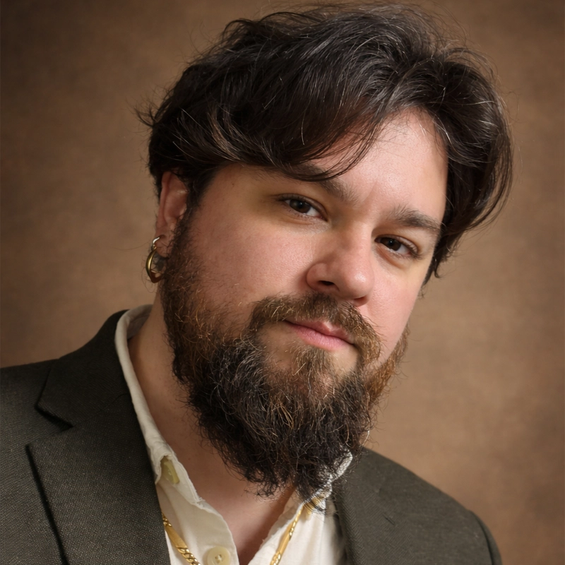 A man with medium-length brown hair and a full beard wears a dark blazer over a white shirt, accessorized with a gold necklace and hoop earring from Rika Sauvage. He poses against a brown backdrop, looking confidently at the camera.