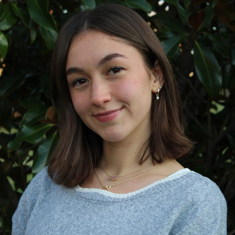 Charlotte Scanlon, a young woman with straight brown hair and light skin, smiles softly in a gray sweater, layered gold necklaces, and small earrings. She stands before lush green foliage, sunlight gently illuminating her face.