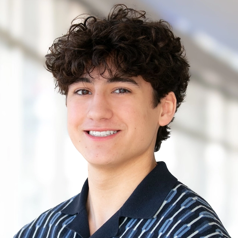 Sophia Leverett, a young person with curly dark hair, smiles at the camera. They wear a dark collared shirt with white stripes. The softly blurred background and bright natural light create a friendly, approachable mood.