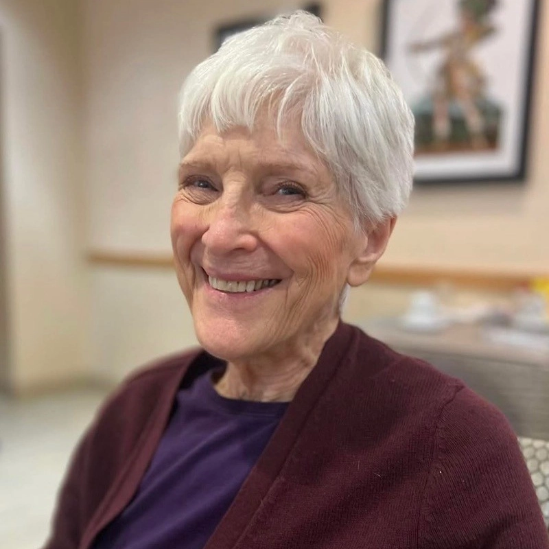 An elderly woman with short white hair smiles warmly at the camera. She is wearing a dark burgundy cardigan over a dark top. The background is softly blurred, showing a framed picture on the wall and a table with dishes.