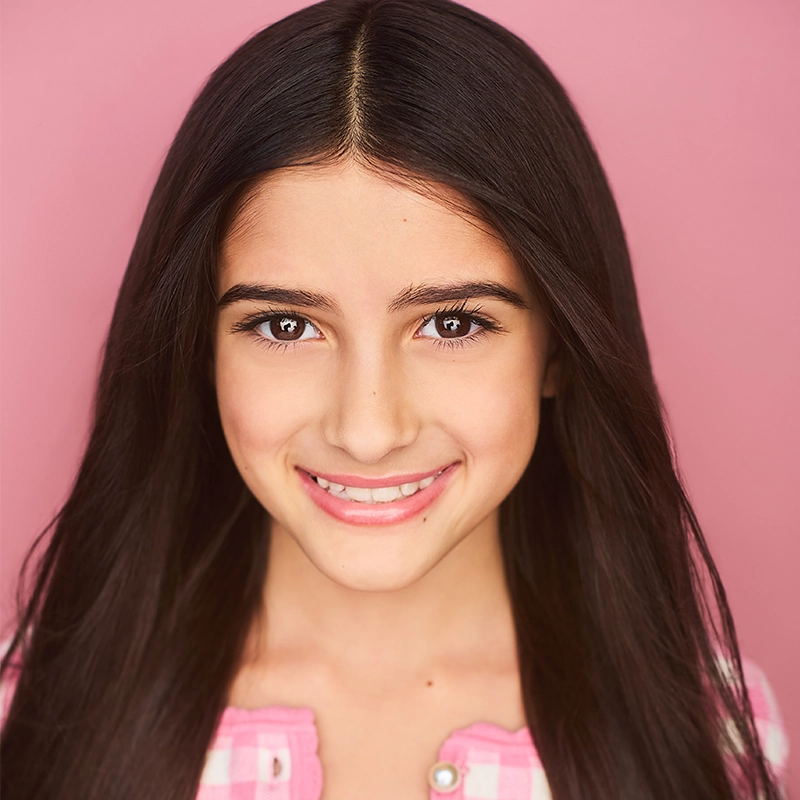 Seraphina Conti, a young person with long, straight, dark hair, smiles warmly at the camera. Wearing a pink and white checkered top against a solid pink background, their expression is friendly and welcoming.