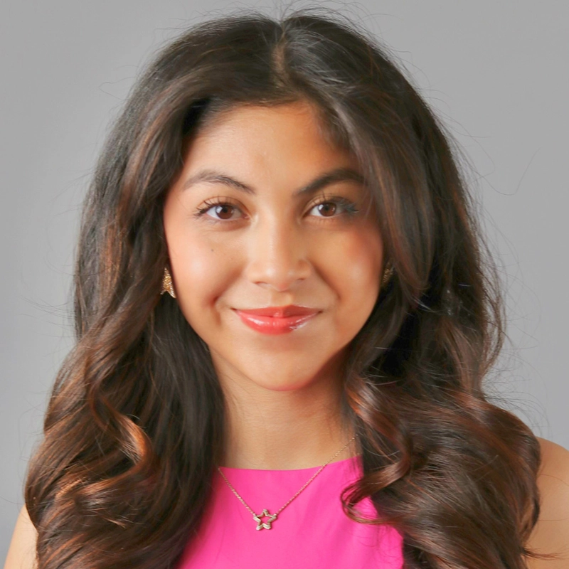 Shayla Lopez, a young woman with long, wavy dark brown hair, smiles softly. She wears a bright pink sleeveless top, gold star necklace, and small gold earrings. Her makeup is natural against a plain light gray background.