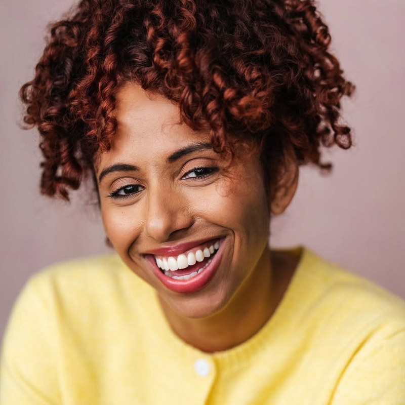 A woman with curly, reddish-brown hair smiles warmly at the camera. She is wearing a yellow sweater and has bright, white teeth. The background is a soft, neutral pinkish color, keeping the focus on her joyful expression.
