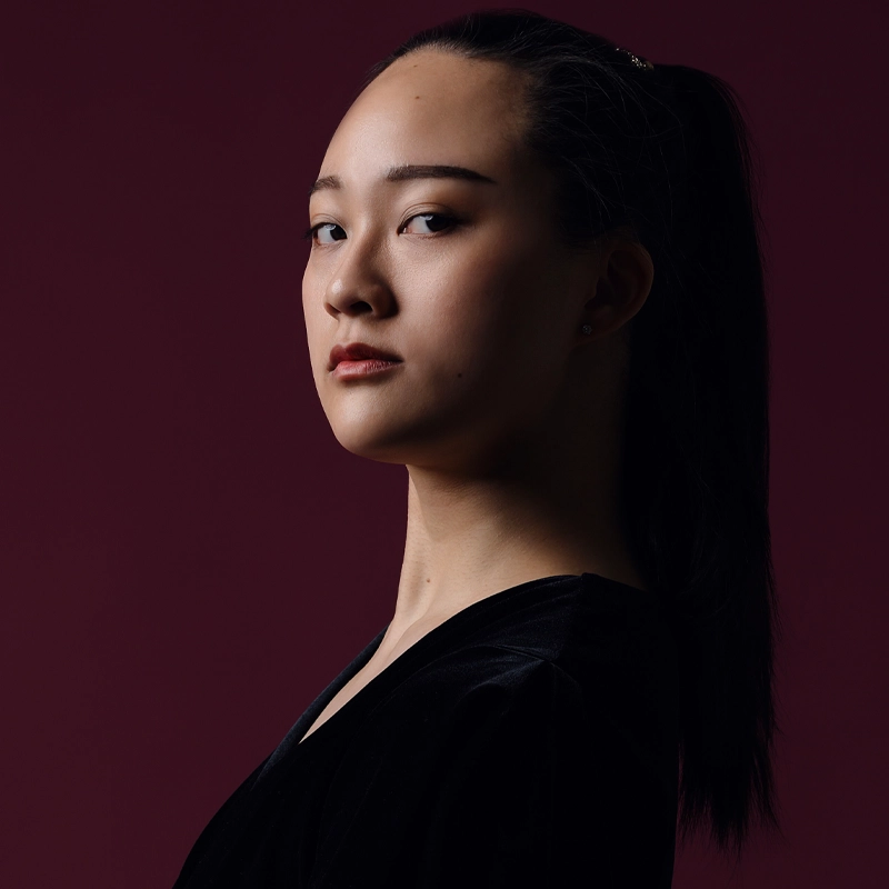 A young woman with long dark hair in a ponytail gazes confidently toward the camera. She wears a black top and poses against a dark maroon background, with dramatic lighting highlighting her face in profile.