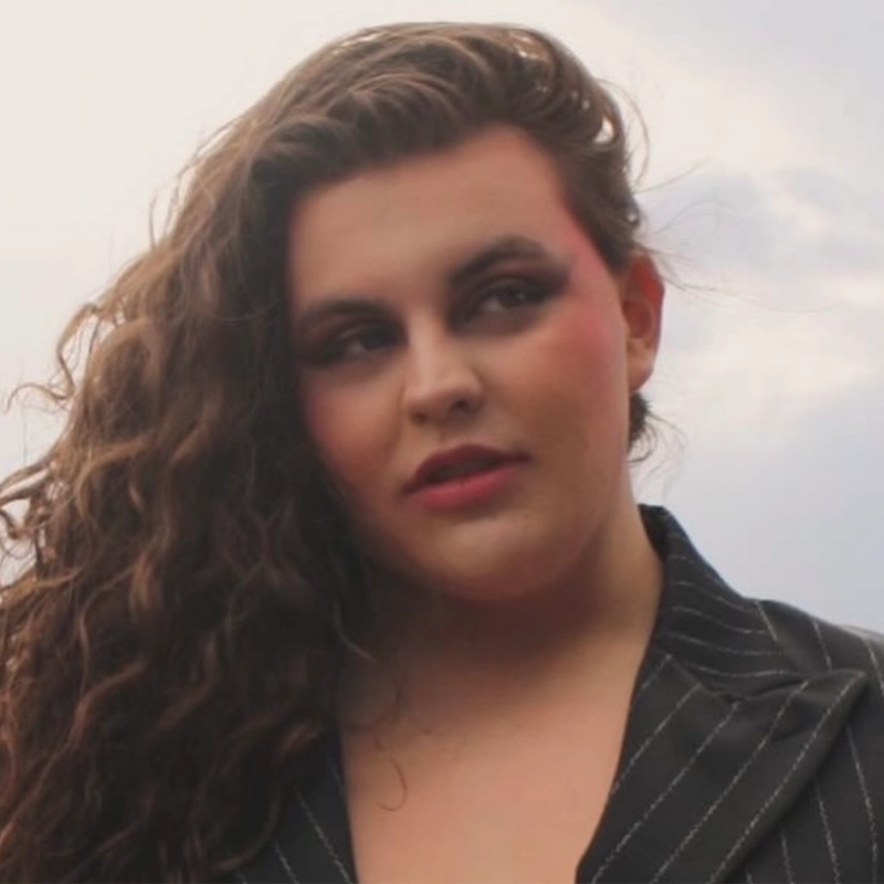 Finch Silverman, with long, wavy brown hair, wears a dark pinstripe blazer and gazes slightly to the side against a cloudy sky. Their bold makeup—red lipstick and blush—complements a calm, thoughtful expression.