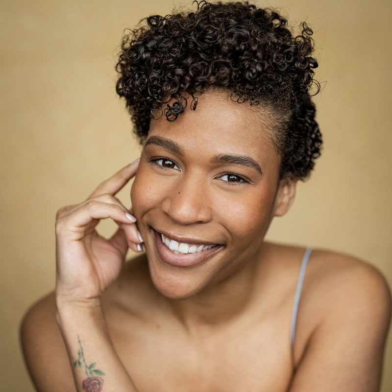 A portrait of Paige Smallwood, a smiling woman with short, curly dark hair. She looks directly at the camera with her hand touching her face, revealing a small rose tattoo on her forearm against a soft, golden-brown background.