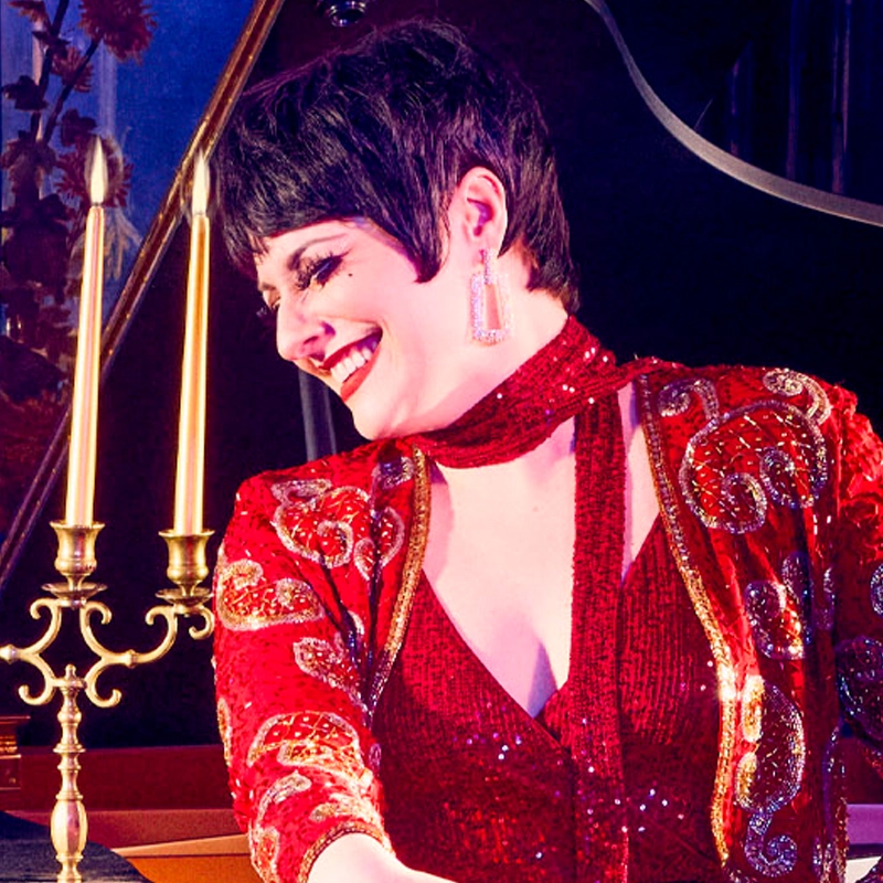 A woman with short dark hair, wearing glittering red attire and large earrings, smiles brightly while looking to the side. Seated by a gold candelabra, Jillian Snow is framed by a dark background and a hint of piano behind her.