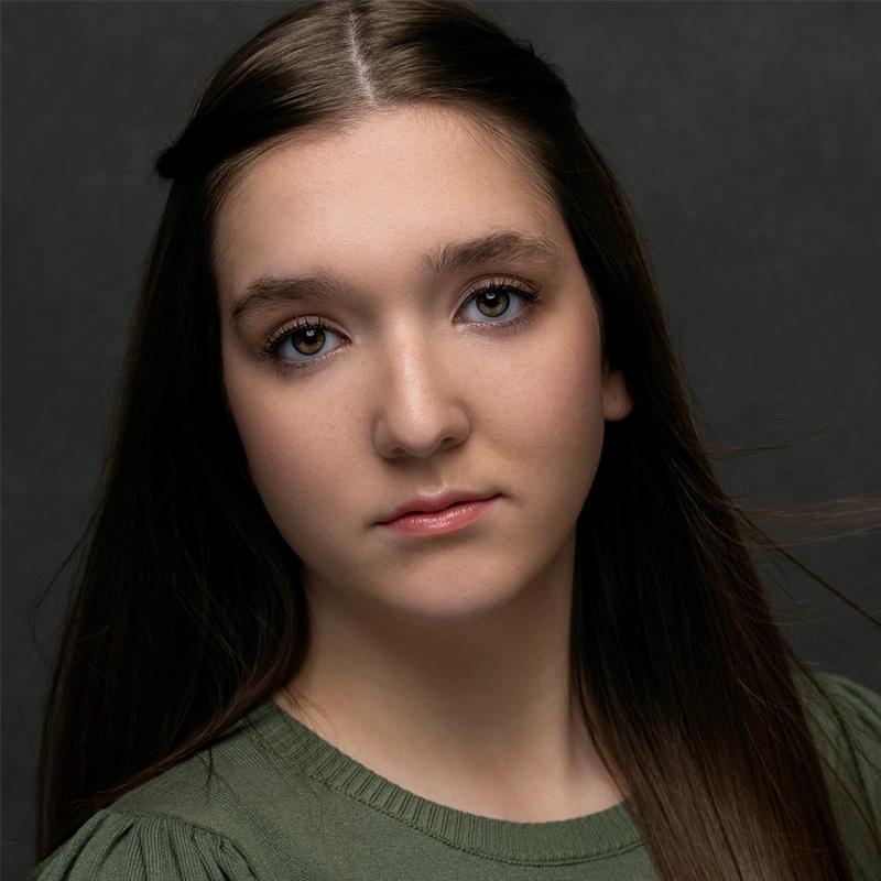 A young woman with fair skin and long brown hair, styled with a middle part, looks directly at the camera. Wearing a green top, Jackie Winslow maintains a neutral expression. The dark, plain background highlights her face.