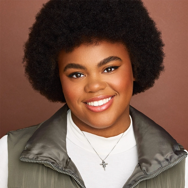 A smiling person with short curly hair wears a puffy gray vest over a white turtleneck. They have neatly groomed eyebrows, warm-toned makeup, and a silver cross necklace. The background is solid brown, featuring Sophia Leverett.